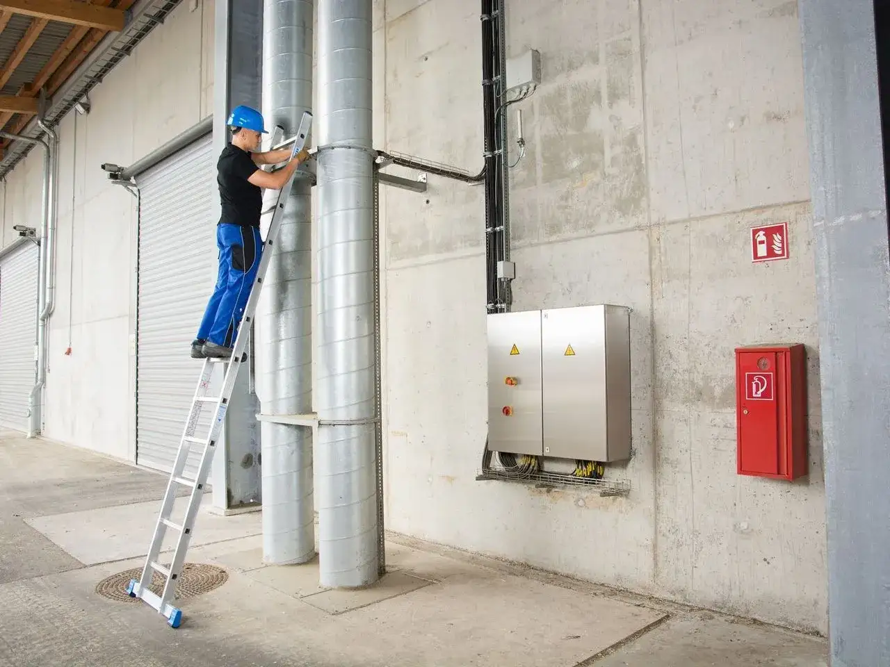 Pracownik na drabinie, kt&oacute;ra jest jednym z wielu rodzaj&oacute;w drabin, sprawdza instalację przy rurach.