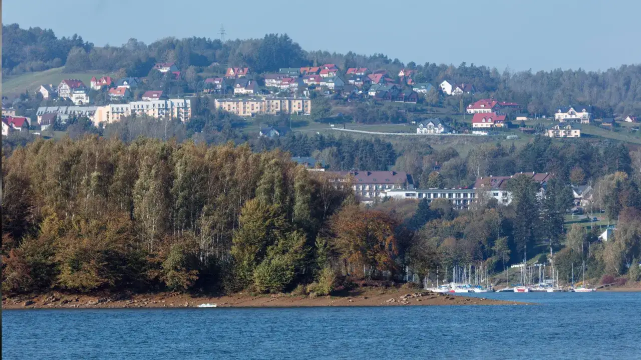 Widok na sanatorium Plon w Polańczyku, z jeziorem i żaglówkami na pierwszym planie, a w tle wzgórza z domami i lasami.