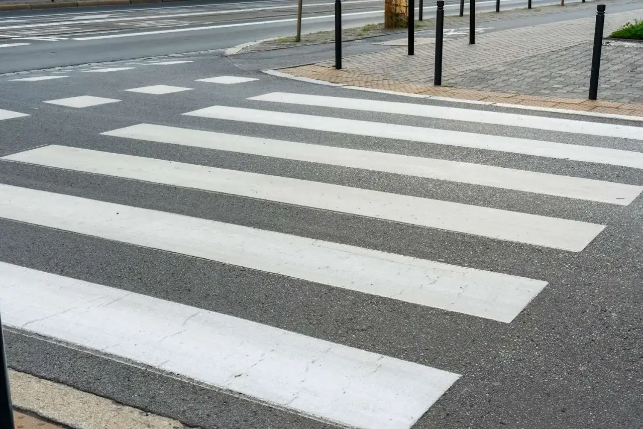 Un passage pi&eacute;ton avec des bandes blanches sur l'asphalte gris. Des poteaux noirs bordent le trottoir pav&eacute;.