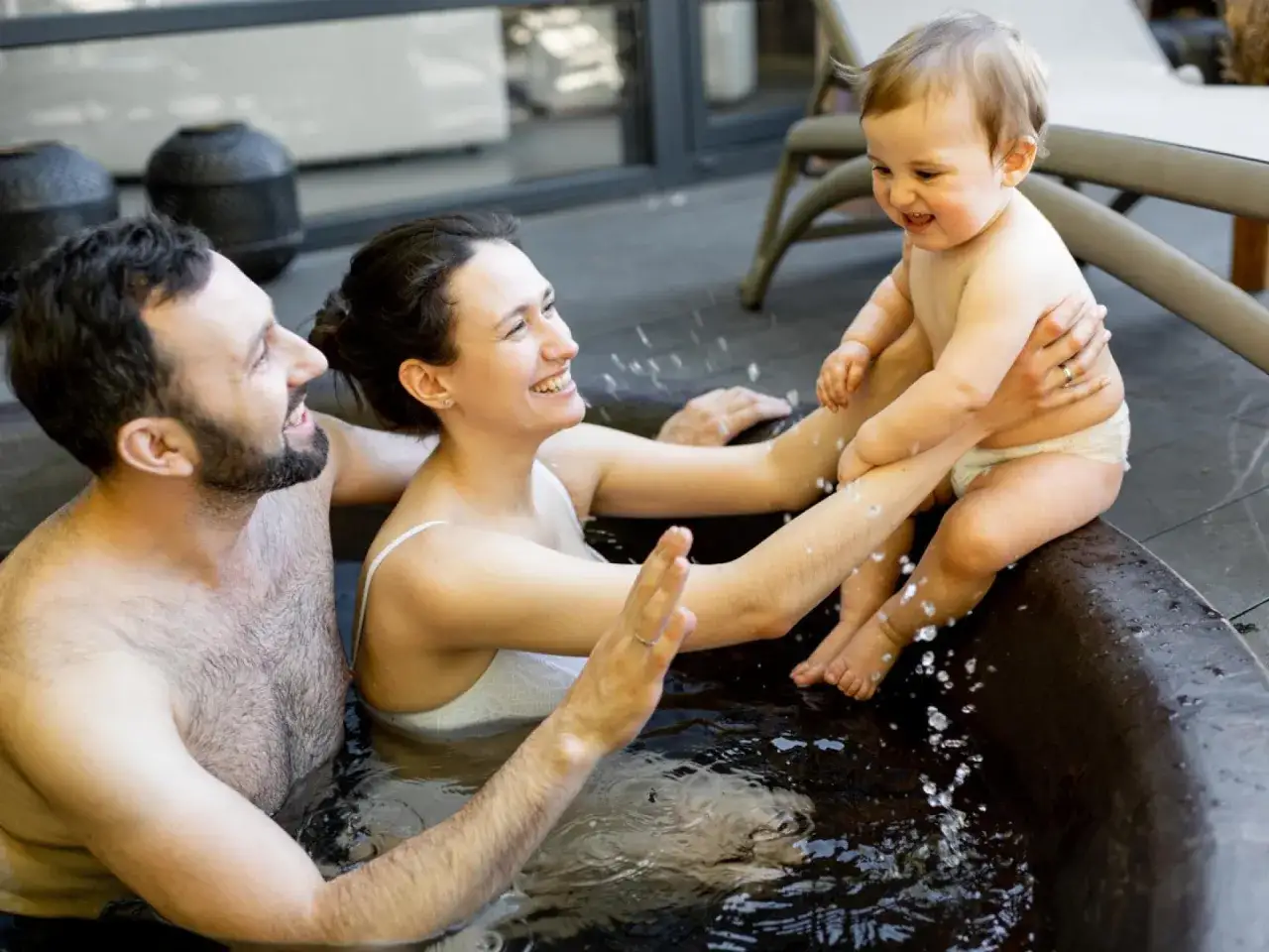 Gl&uuml;ckliche Familie mit 8 Wochen altem Baby im Schwimmbad. Das Baby planscht fr&ouml;hlich im Wasser, w&auml;hrend die Eltern es liebevoll halten.