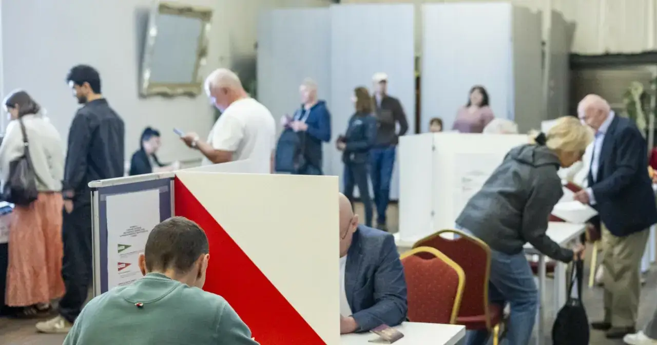 Ludzie głosują w lokalu wyborczym za granicą. Widać kabiny do głosowania i osoby czekające w kolejce.