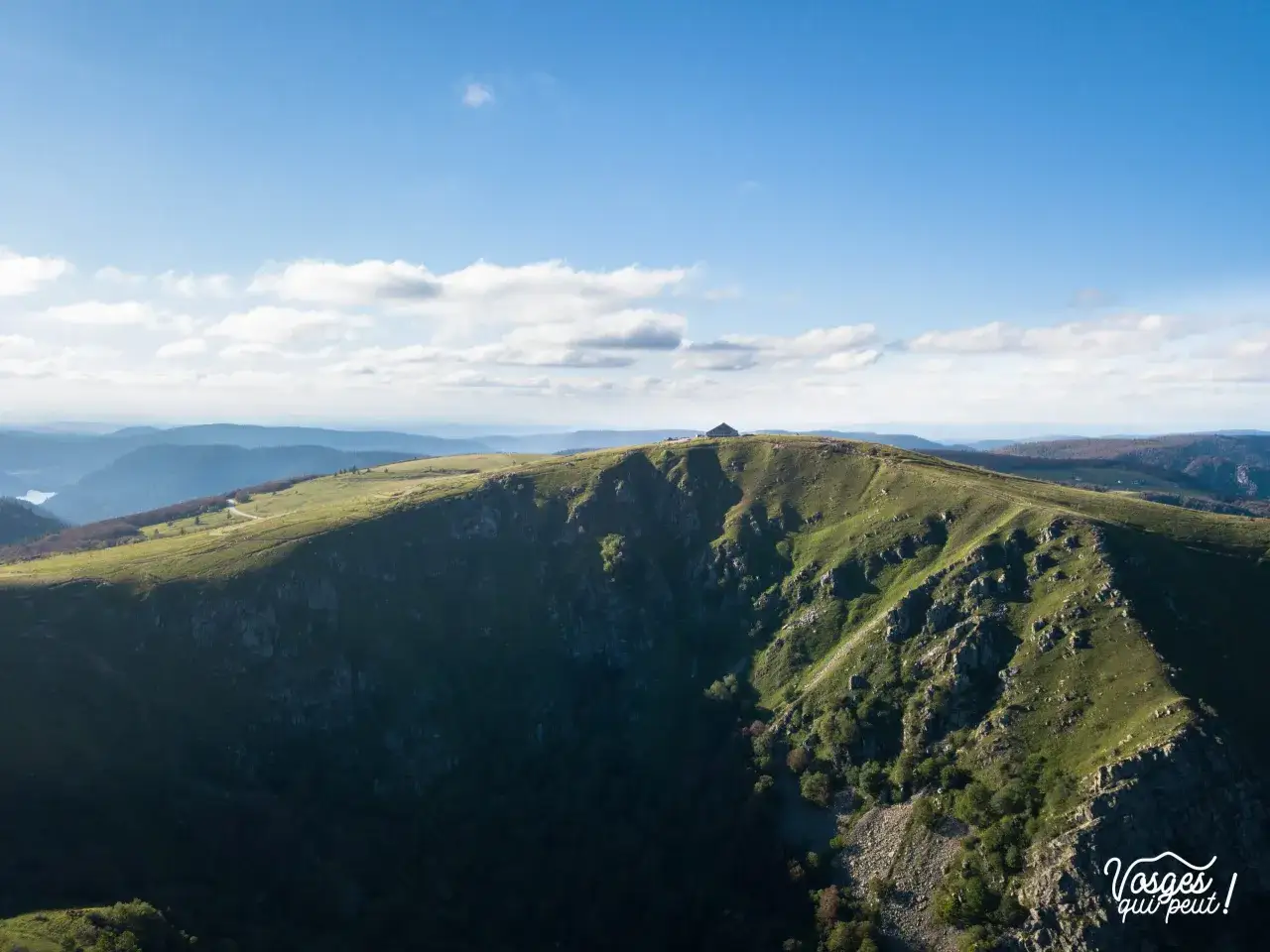 Vue panoramique des **Vosges** sous un ciel bleu et nuageux. Un sentier serpente sur une cr&ecirc;te verdoyante menant &agrave; un refuge.