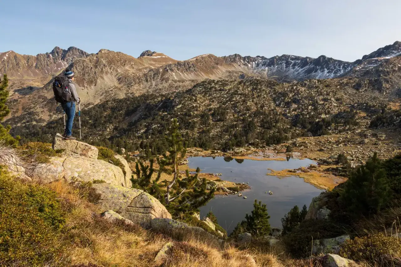 Randonnée en Andorre : un marcheur admire un lac de montagne entouré de rochers et de pins, avec des sommets enneigés en arrière-plan.