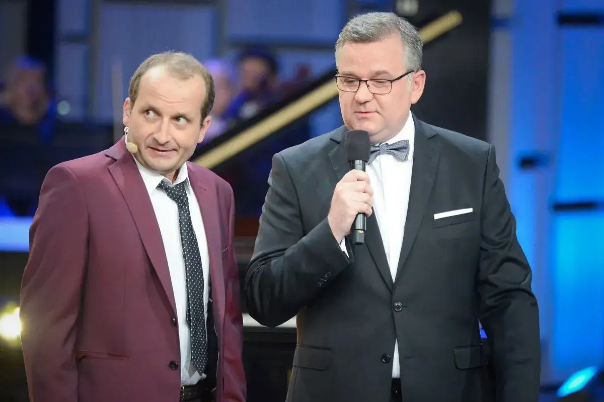Dwóch mężczyzn w garniturach, jeden z nich trzyma mikrofon. To prawdopodobnie szkło kontaktowe obsada.