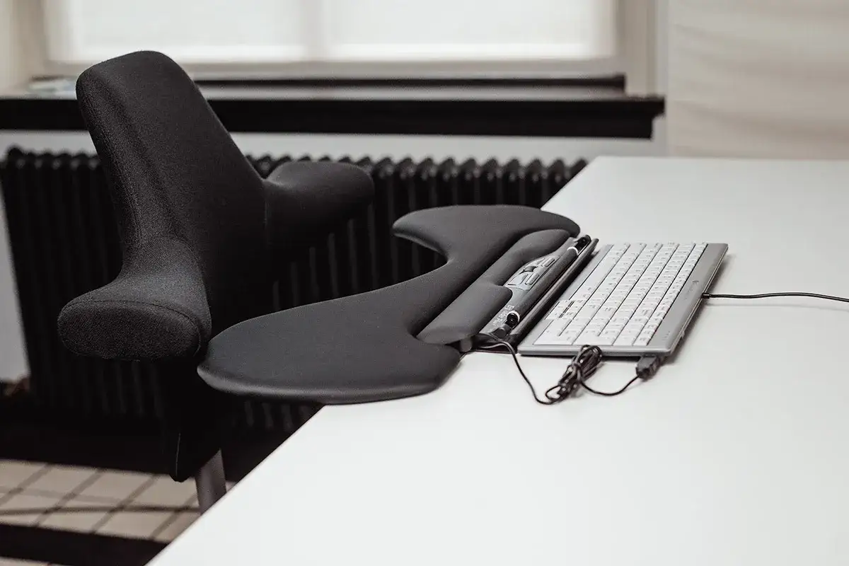 Ergonomiskt stöd för mus arm och tangentbord på ett vitt skrivbord. En svart stol syns till vänster.