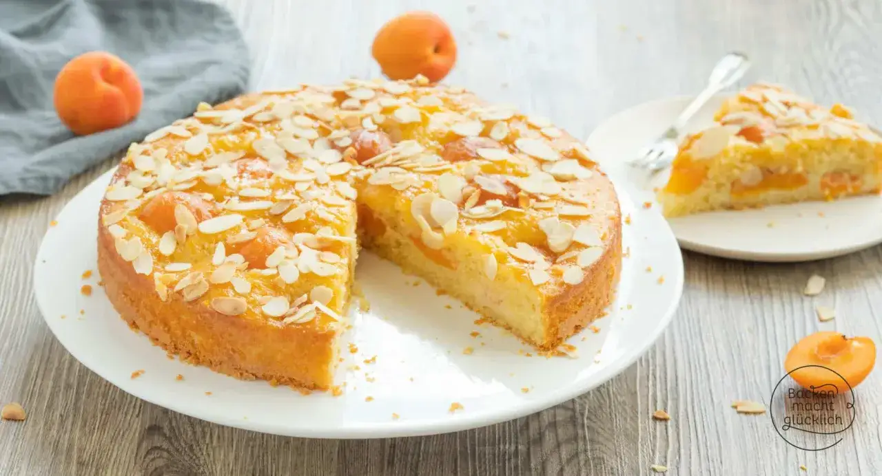 Saftiger aprikosenkuchen mit mandeln, garniert mit frischen Aprikosen und Mandelbl&auml;ttchen. Ein St&uuml;ck ist bereits auf einem Teller angerichtet.
