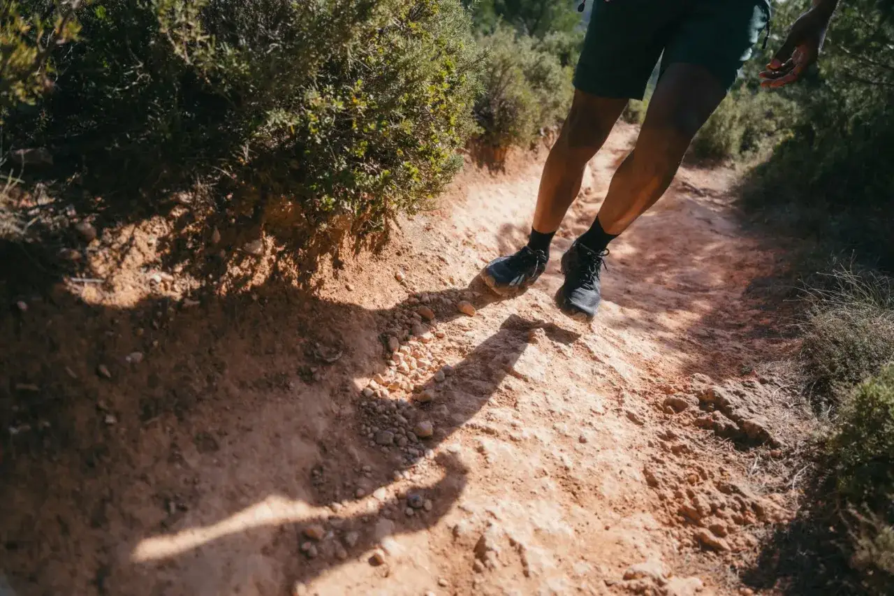 Jambes d'un coureur en plein fast hiking sur un sentier rocailleux et poussiéreux, entouré de végétation sèche.