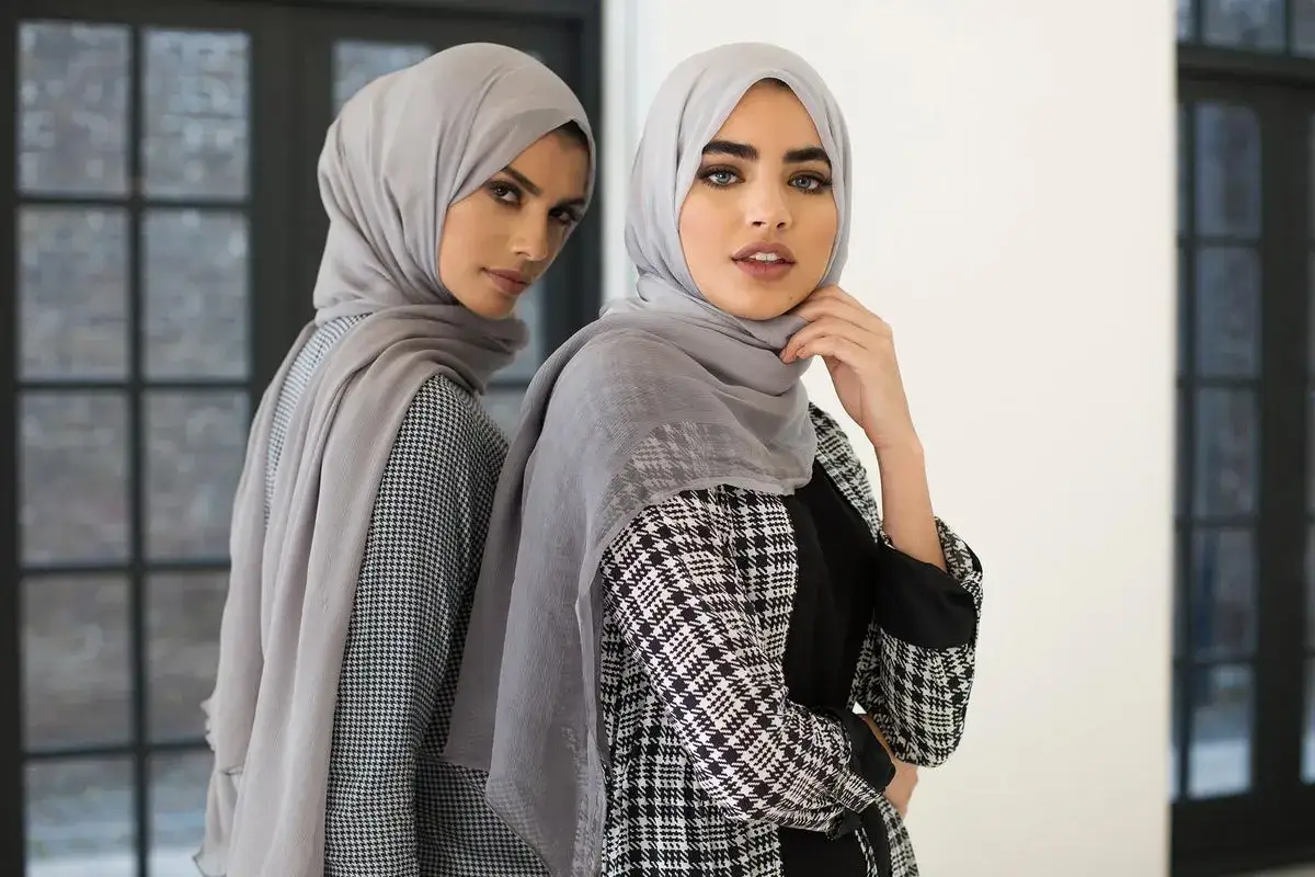 Two women showcase modest clothing, wearing grey hijabs and stylish patterned abayas.