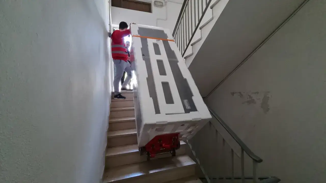 Zwei M&auml;nner transportieren einen gro&szlig;en, verpackten Trockner &uuml;ber eine Treppe. Das Gewicht eines Trockners kann variieren.