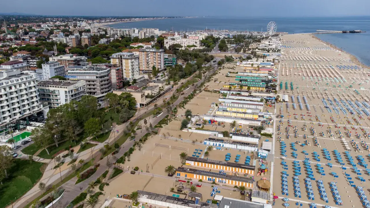 Vista aerea di Rimini spiaggia libera con file ordinate di ombrelloni e cabine colorate, hotel e mare all'orizzonte.