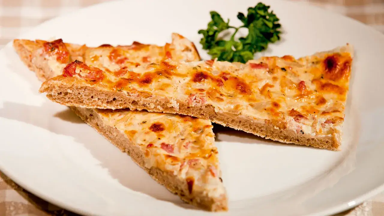 Drei St&uuml;cke eines herzhaften Zwiebelkuchens, ein klassisches Ploatz-Rezept, auf einem wei&szlig;en Teller mit Petersilie garniert.