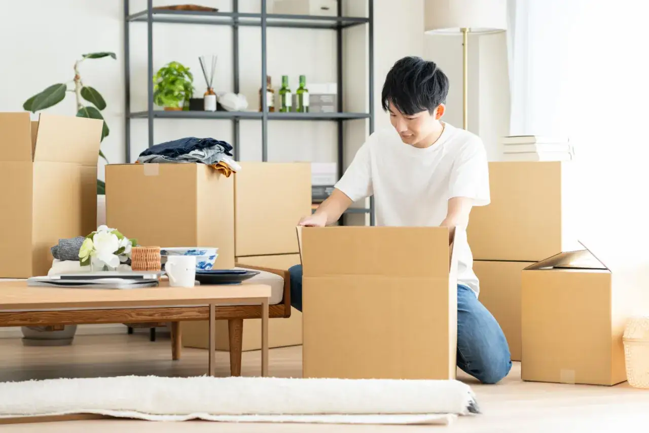 Ein Mann packt Kartons ein, um sie aufzubewahren. Umzugskartons und Haushaltsgegenst&auml;nde sind auf dem Boden und einem Tisch verteilt.