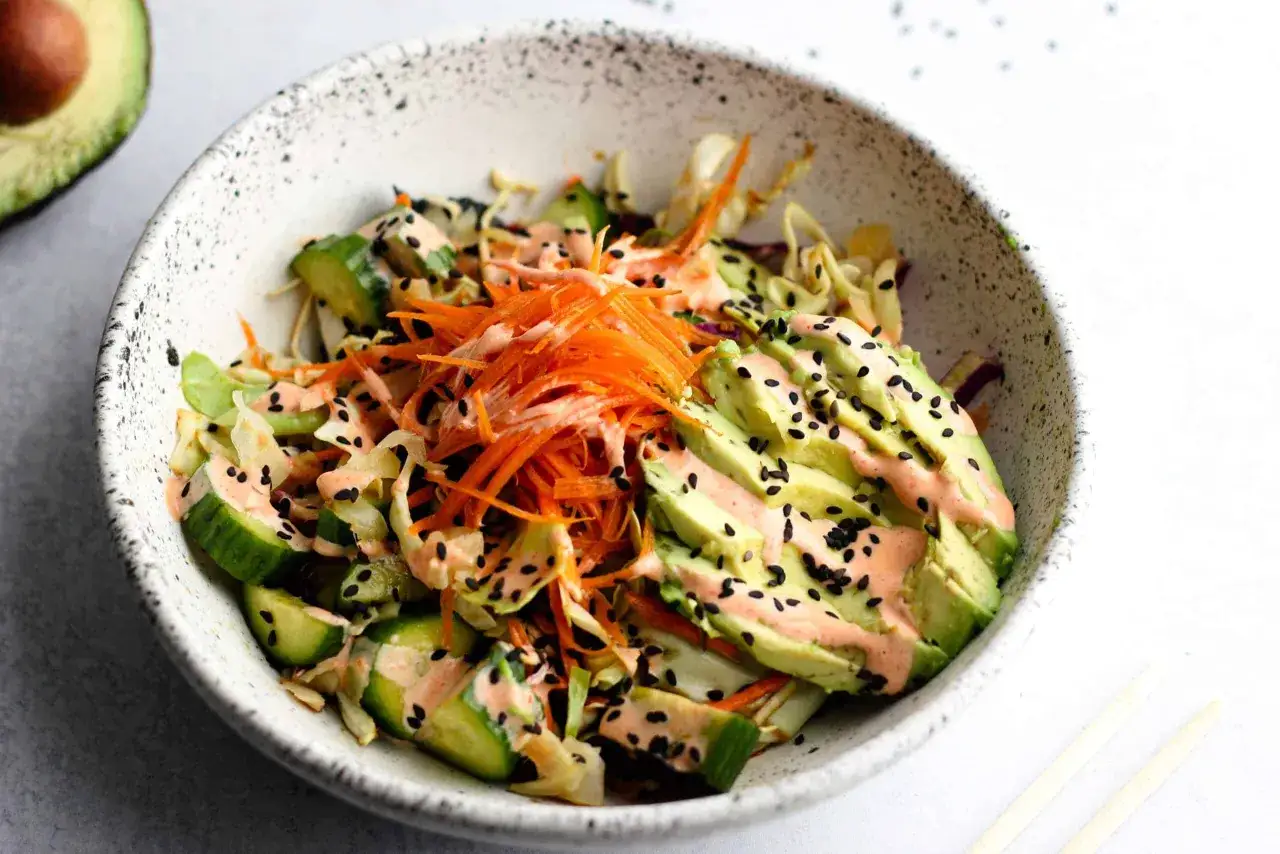 Schüssel mit asiatischem Salat: Avocado, Gurke, Karottenstreifen, Kohl und Sesam-Dressing, bestreut mit schwarzen Sesamkörnern.