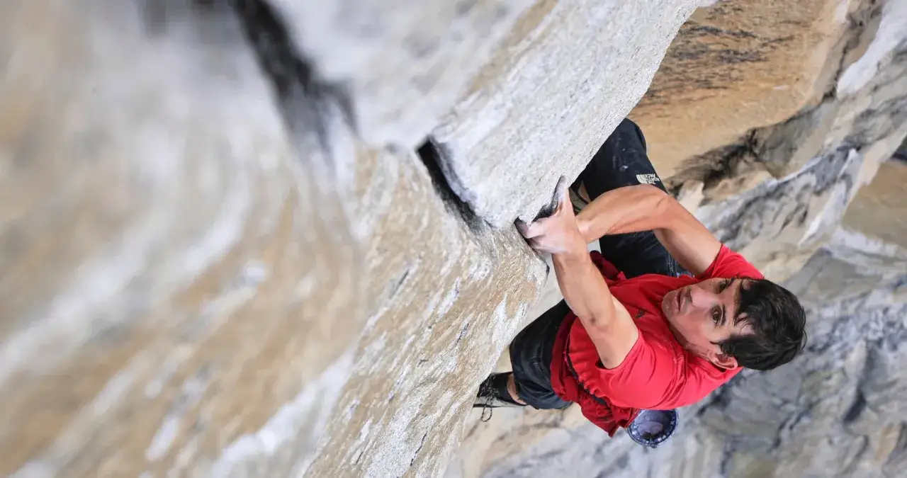 Un escalador con camiseta roja se aferra a una pared de roca, capturado en un momento de tensión para un documental de escalada.