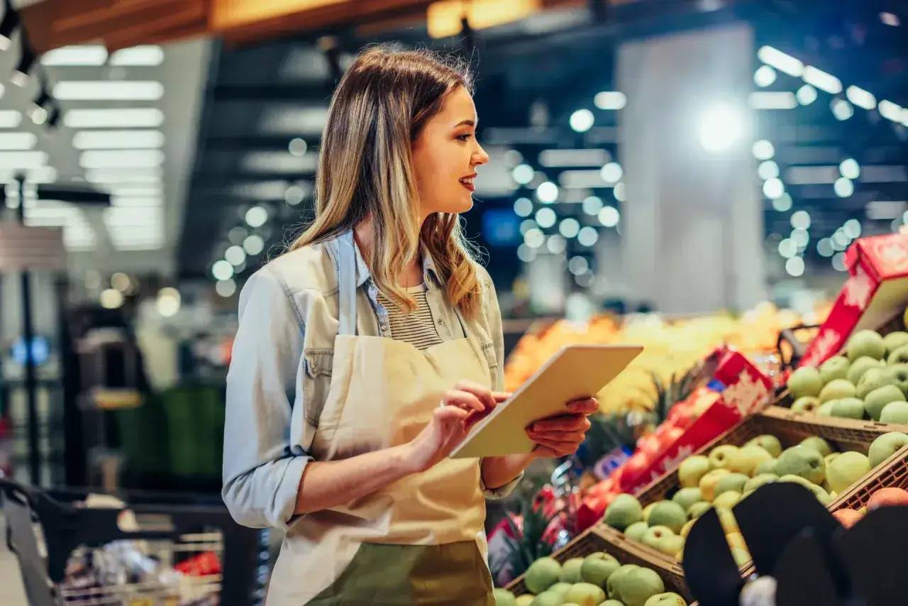 Pracownica Żabki z tabletem w ręku, zastanawia się, ile zarabia pracownik Żabki netto, przeglądając dane o sprzedaży owoców.