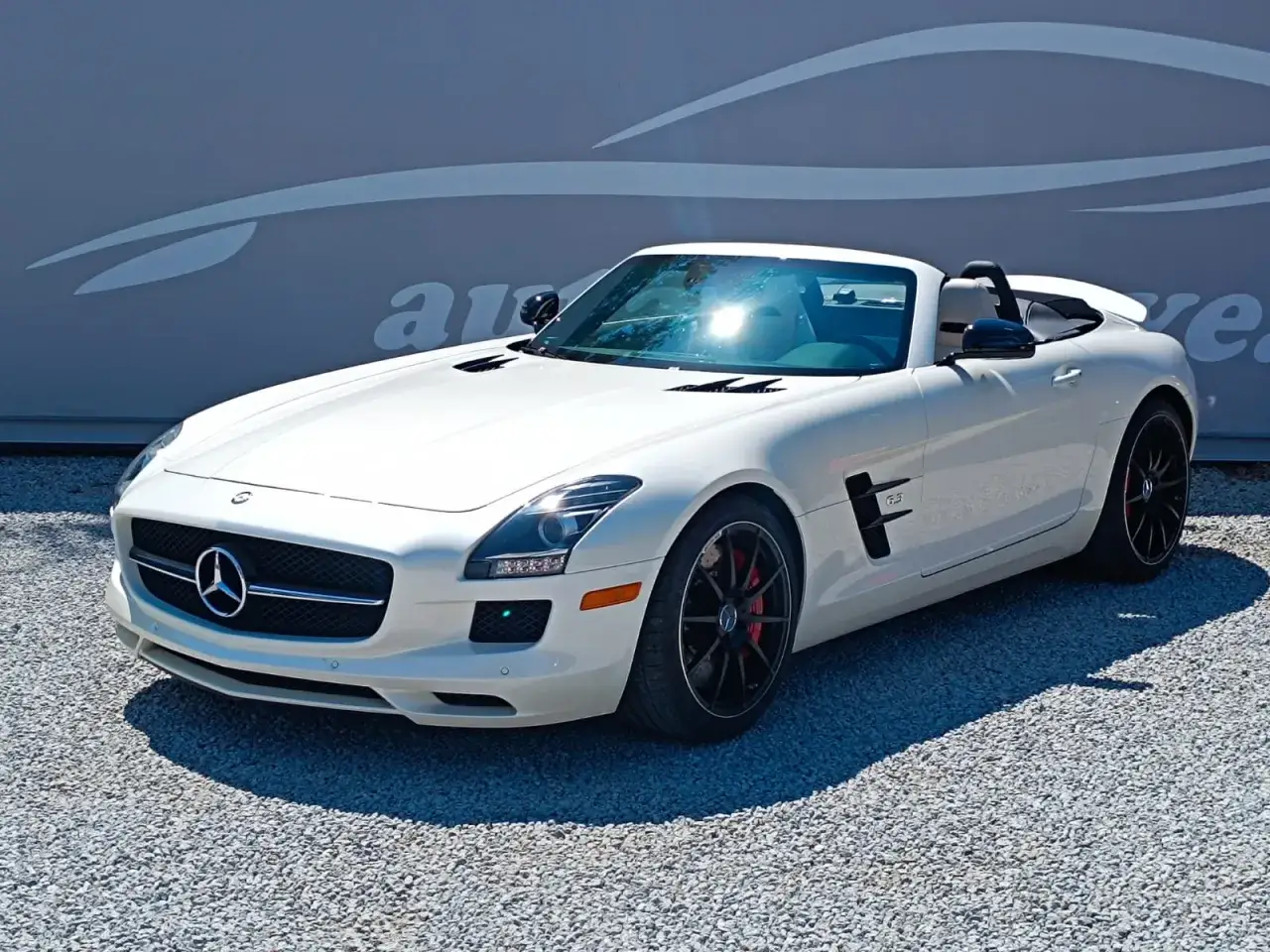 Biały kabriolet SLS AMG z czarnymi felgami i czerwonymi zaciskami hamulcowymi, zaparkowany na żwirze.