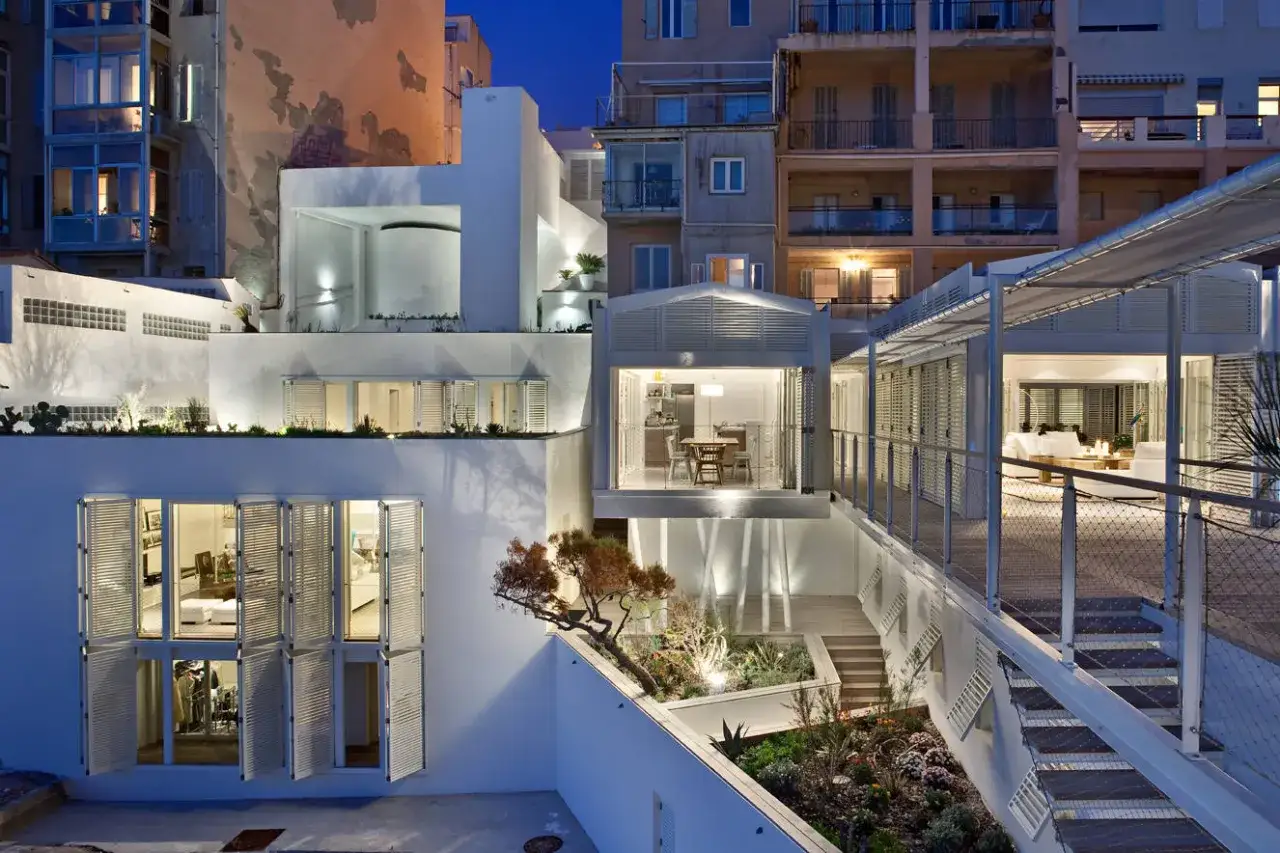 Une maison moderne aux volets blancs ouverts, illuminée la nuit. Un photographe architecture capture la beauté de ce lieu.