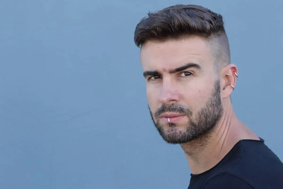 Mann mit Undercut und Bart, Lippen- und Ohrpiercings vor blauem Hintergrund.