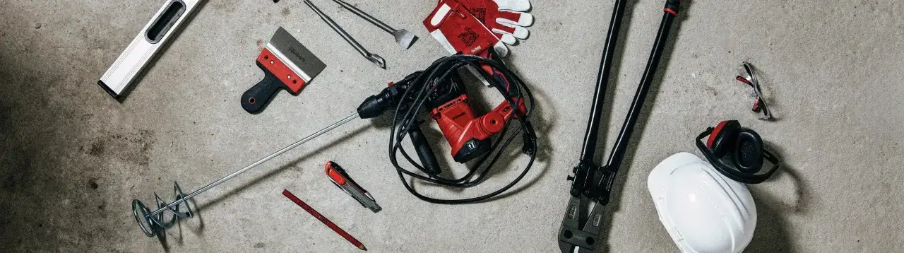 Narzędzia na budowę: mieszadło, młotowiertarka, rękawice, poziomica, nożyk, ołówek, nożyce do prętów, kask, okulary i słuchawki ochronne.