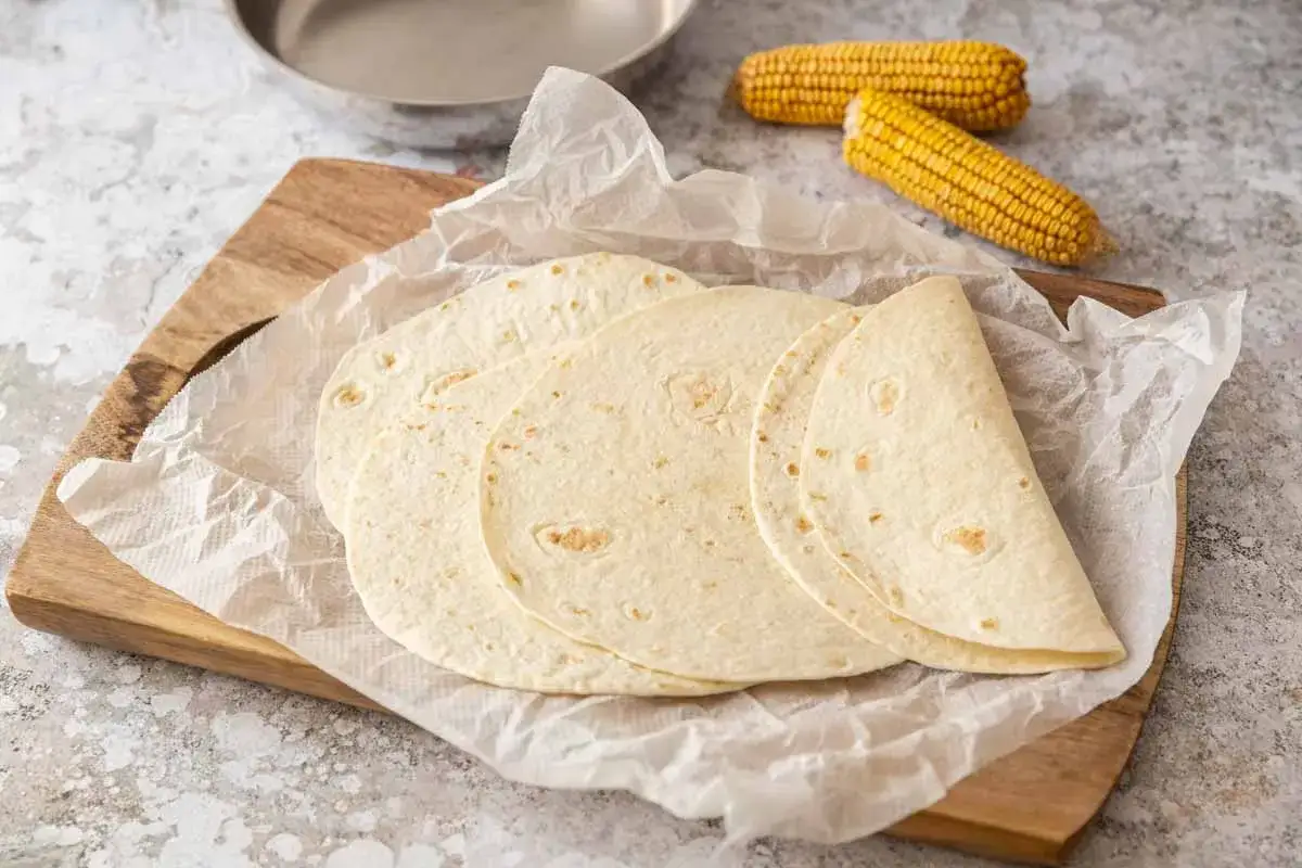 Des tortillas maison pr&ecirc;tes &agrave; &ecirc;tre garnies, avec des &eacute;pis de ma&iuml;s en arri&egrave;re-plan. Parfait pour une recette facile et savoureuse.