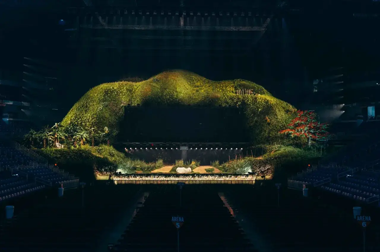 Scenografia da concerto con una collina verde lussureggiante, palme e un albero rosso. Sembra una 