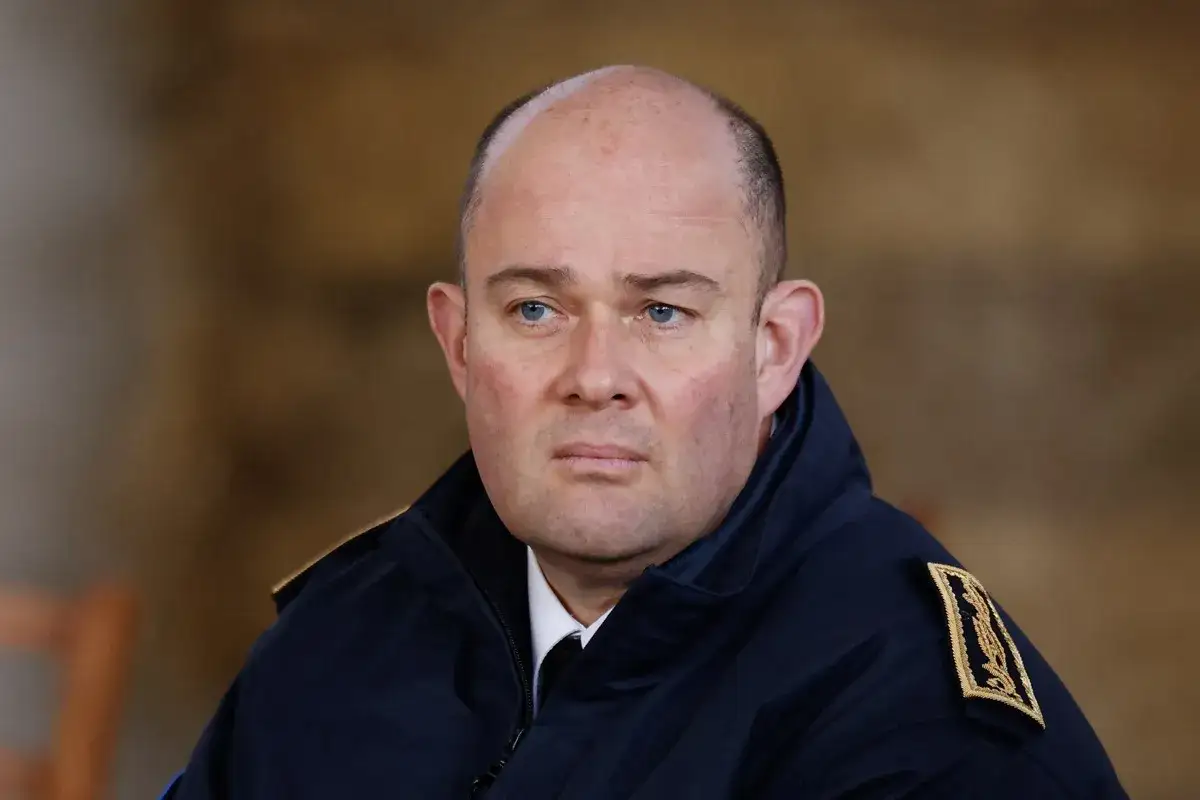 Un homme chauve aux yeux bleus, portant un uniforme bleu marine avec des épaulettes dorées, ressemble à un chef d'état-major en mission.