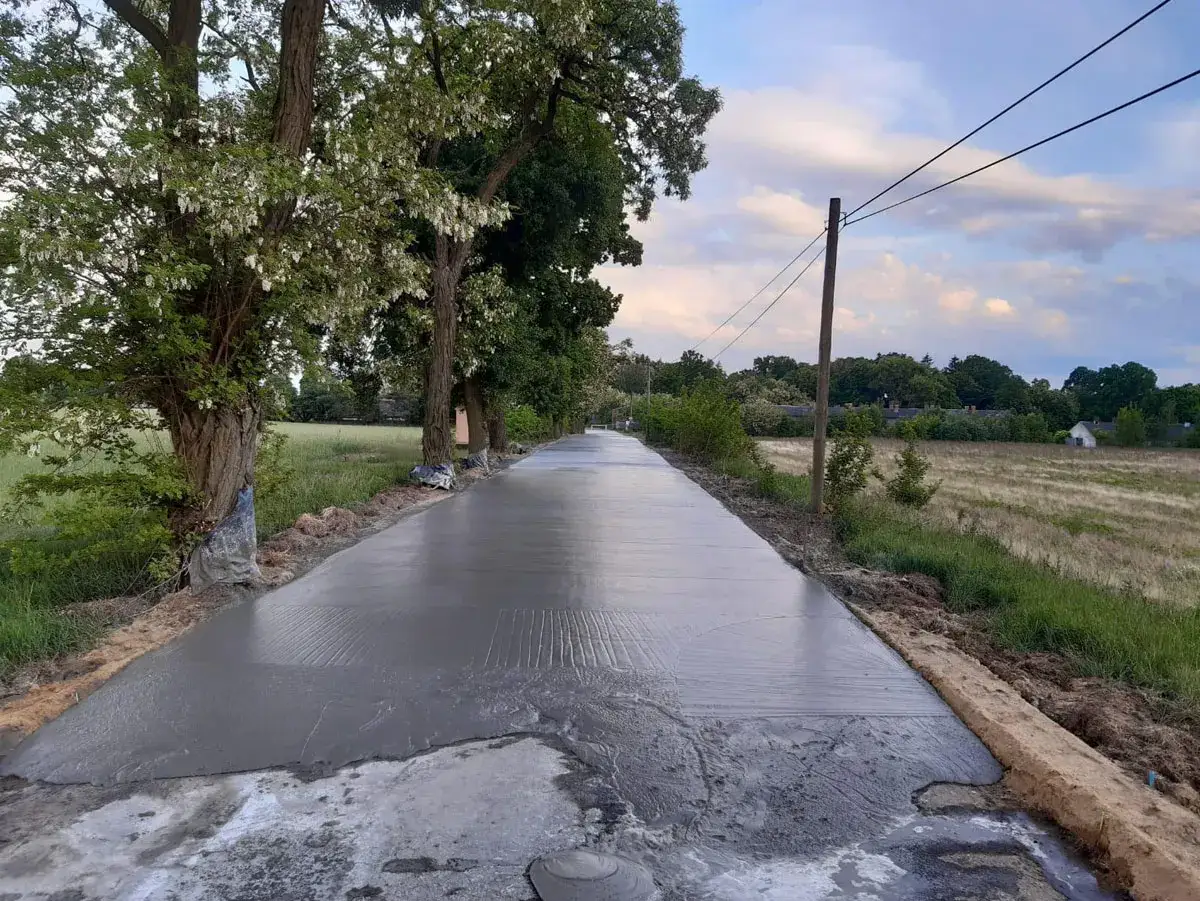 Świeżo wylany beton na drodze, otoczonej drzewami i polami. Zastanawiasz się, ile kosztuje metr sześcienny asfaltu?