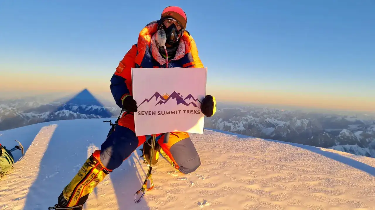 Kristin Harila auf dem K2, h&auml;lt ein Schild von Seven Summit Treks. Der Schatten des Berges wirft sich in die Ferne.