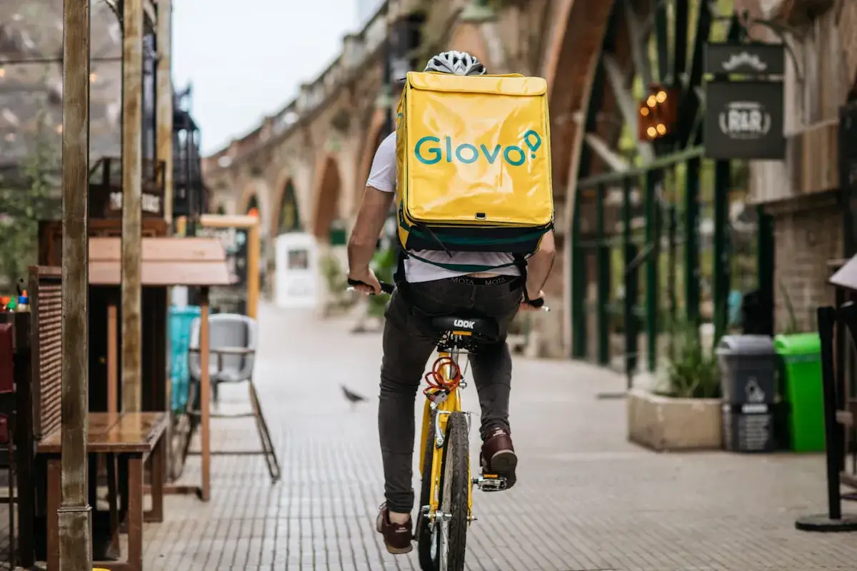 Kurier Glovo na rowerze z ż&oacute;łtym plecakiem przemierza ulicę. Szybka dostawa z aplikacji lisek.