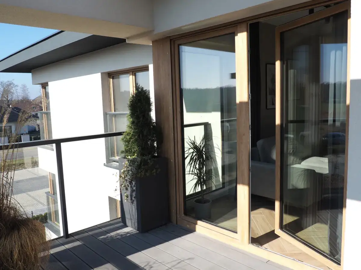 Nowoczesny balkon z drewnianymi, podw&oacute;jnymi drzwiami tarasowymi. Okna dzielone poziomo wpuszczają dużo światła.