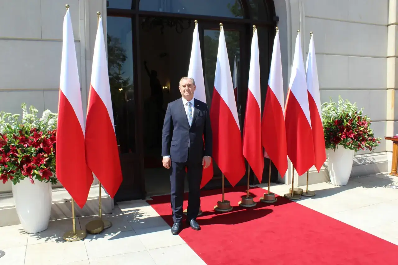 Mężczyzna w garniturze stoi przed polskimi flagami. Czy to burmistrz, czy prezydent? Trudno powiedzieć, ale wygląda uroczyście.