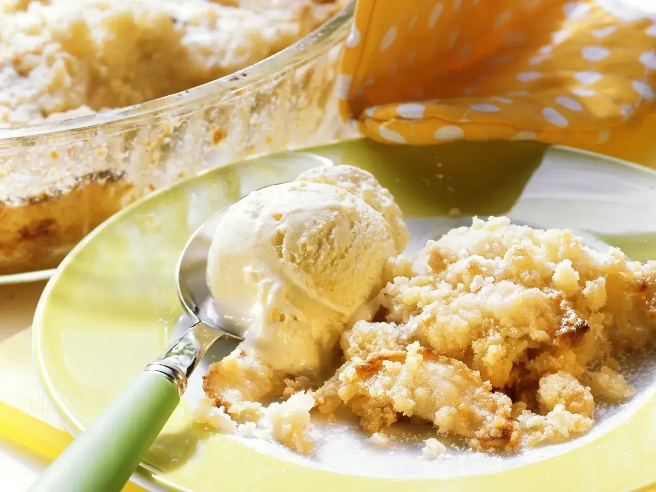 Ein Teller mit warmem Apple Crumble, bestreut mit Puderzucker, daneben eine Kugel Vanilleeis auf einem L&ouml;ffel.