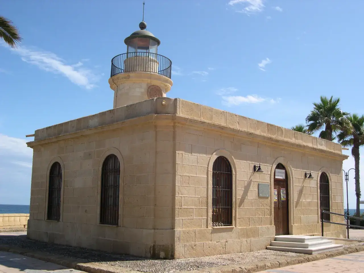 El faro del Sabinar, con su torre circular y edificio anexo de piedra, se alza bajo un cielo azul. Palmeras se ven al fondo.