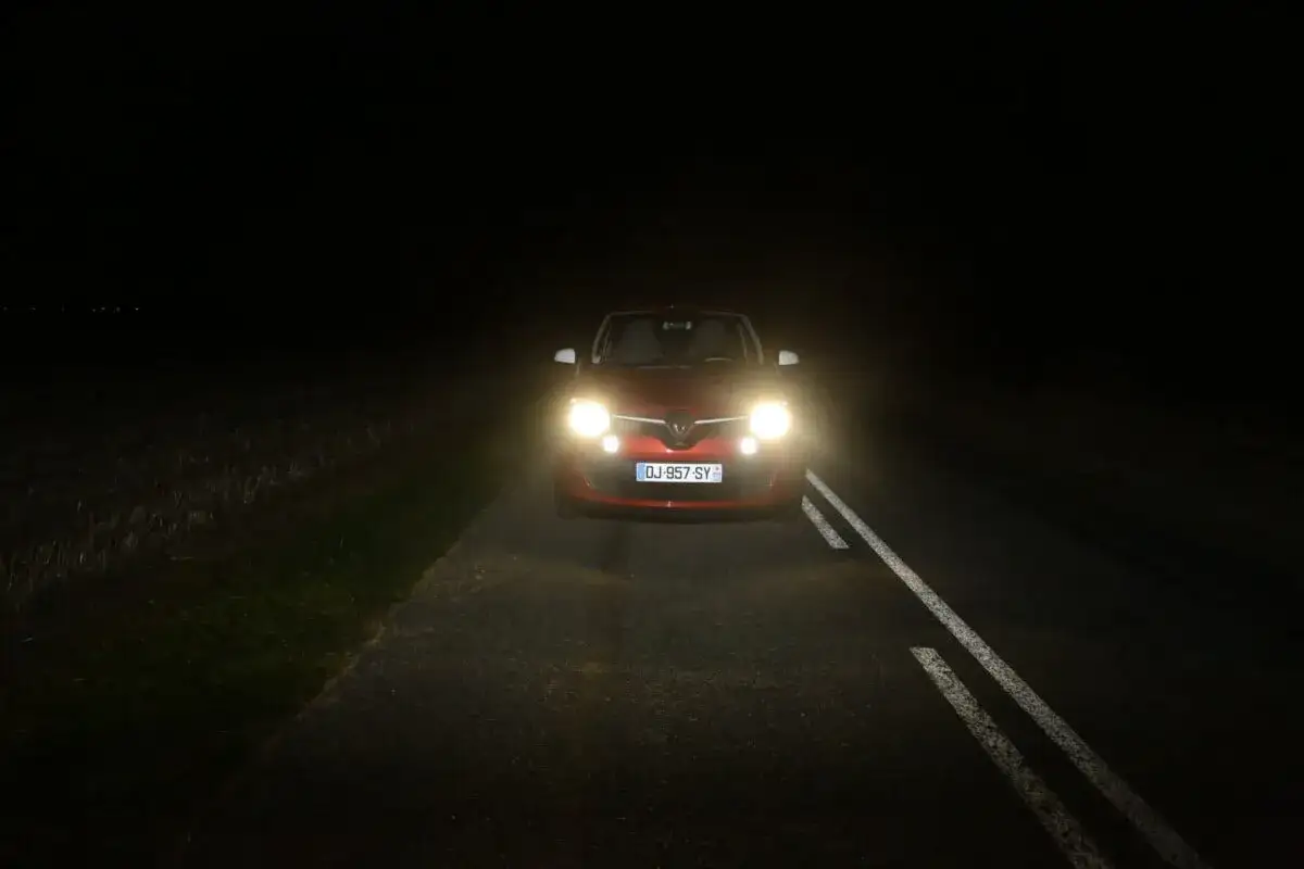 Une voiture rouge roule sur une route sombre, ses feux de route éclairent le chemin. Quand les utiliser ? La nuit, sur route dégagée, pour une meilleure visibilité.