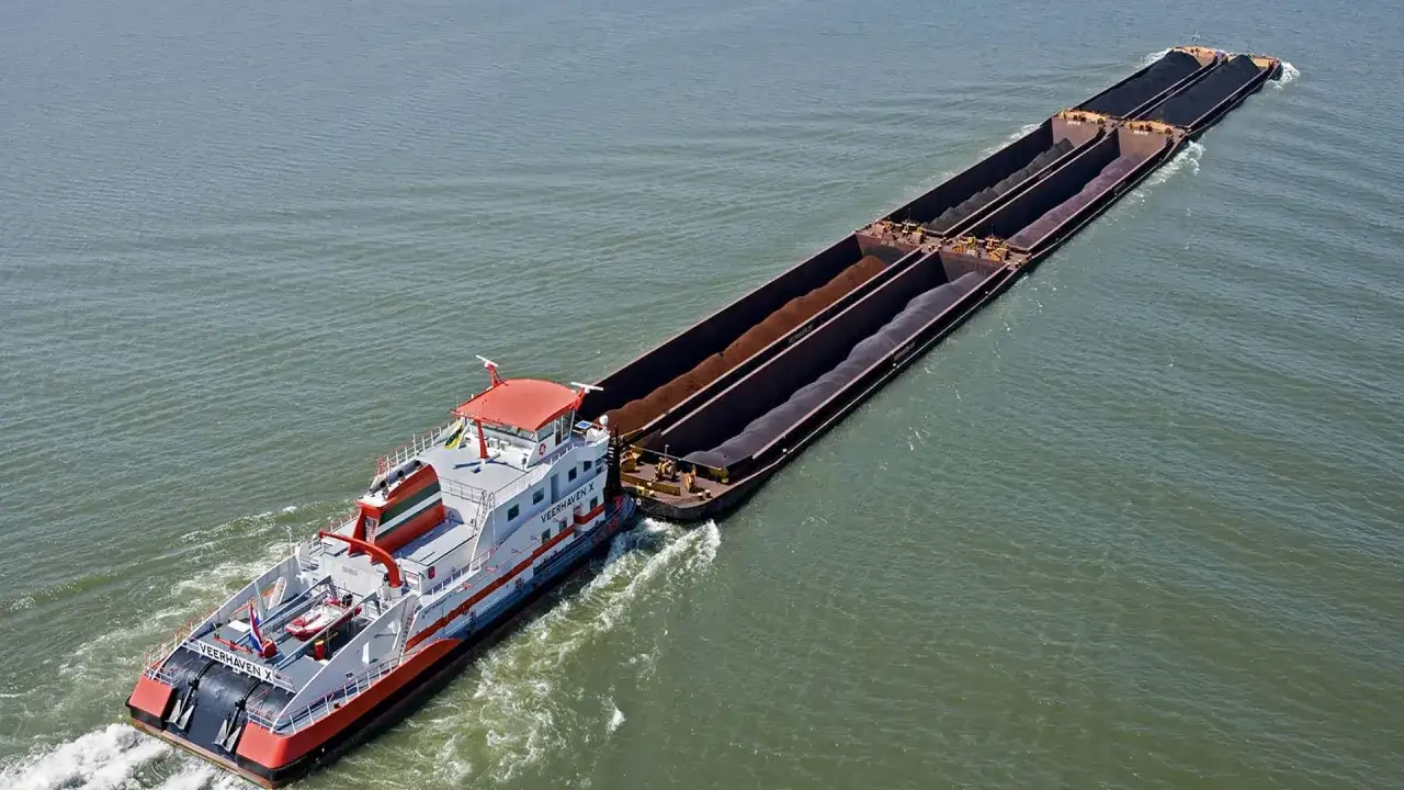 Ein Schlepper zieht ein langes barge ship mit Kohle beladen über das Wasser.