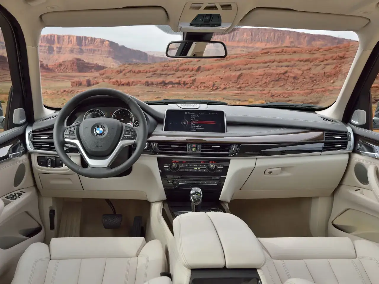 Wnętrze BMW X5, widok z perspektywy kierowcy. Na desce rozdzielczej ekran systemu multimedialnego, kierownica z logo BMW. Za oknem pustynny krajobraz.