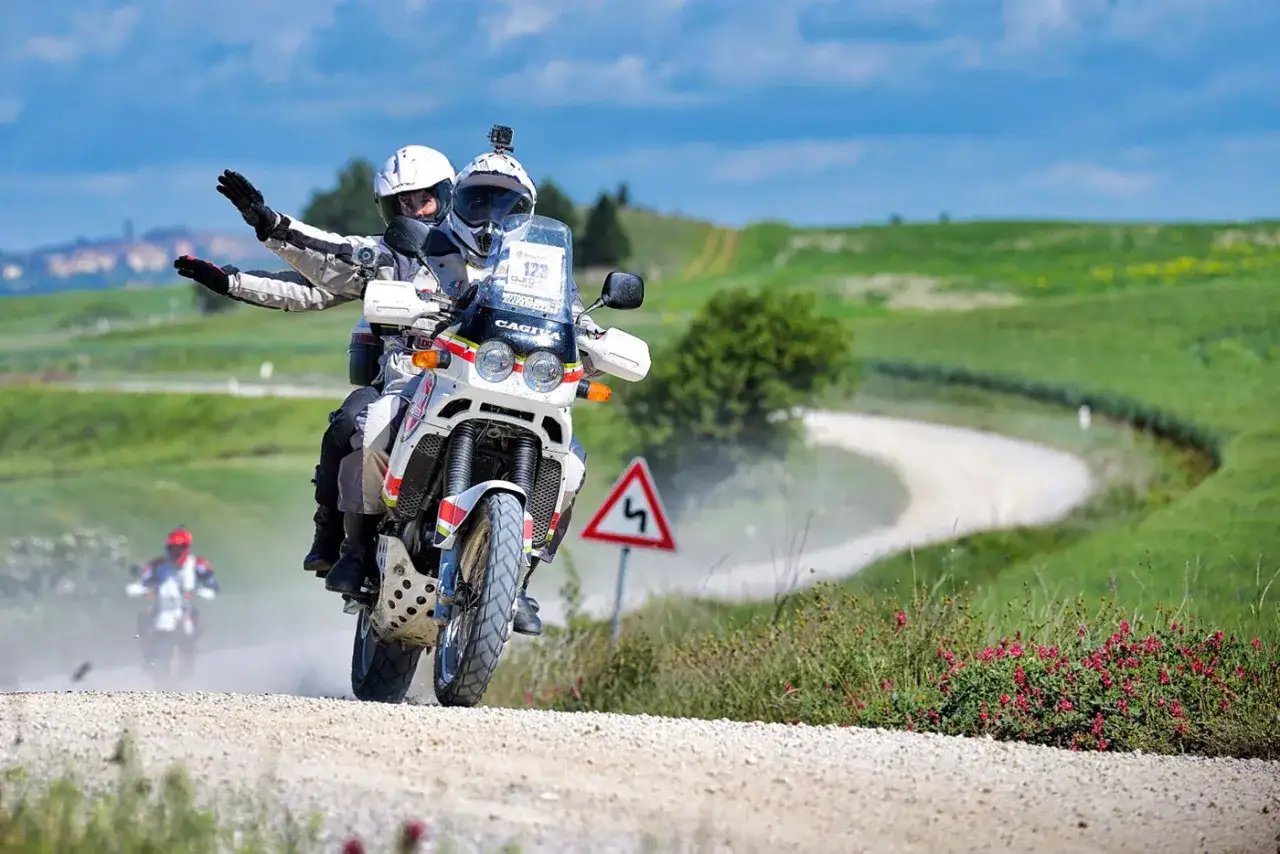 Coppia in moto affronta un percorso eroico in moto su sterrato, tra colline verdi e cielo azzurro.