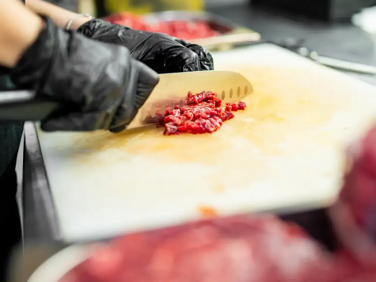 Ręce w czarnych rękawiczkach siekają mięso. Dbałość o higienę zapobiega rozwojowi bakterii, takich jak listeria.