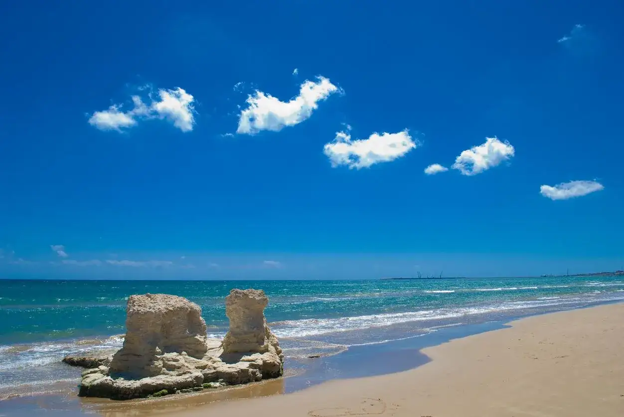 Scogliere calcaree sulla spiaggia dorata, mare azzurro e cielo limpido in **Sicilia orientale**.