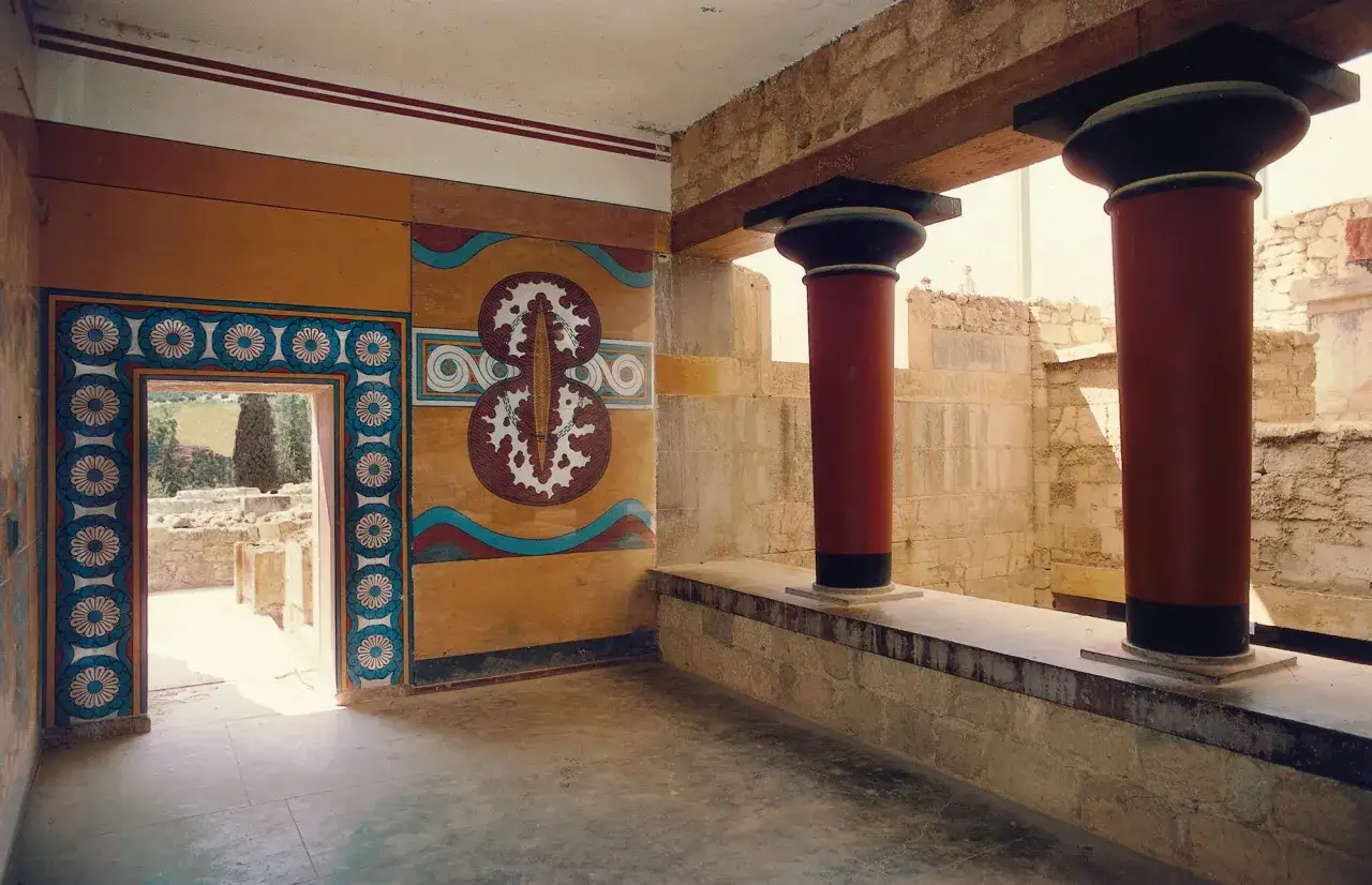 Interior del palacio de Cnosos con columnas rojas, murales coloridos y una puerta que da a ruinas antiguas.