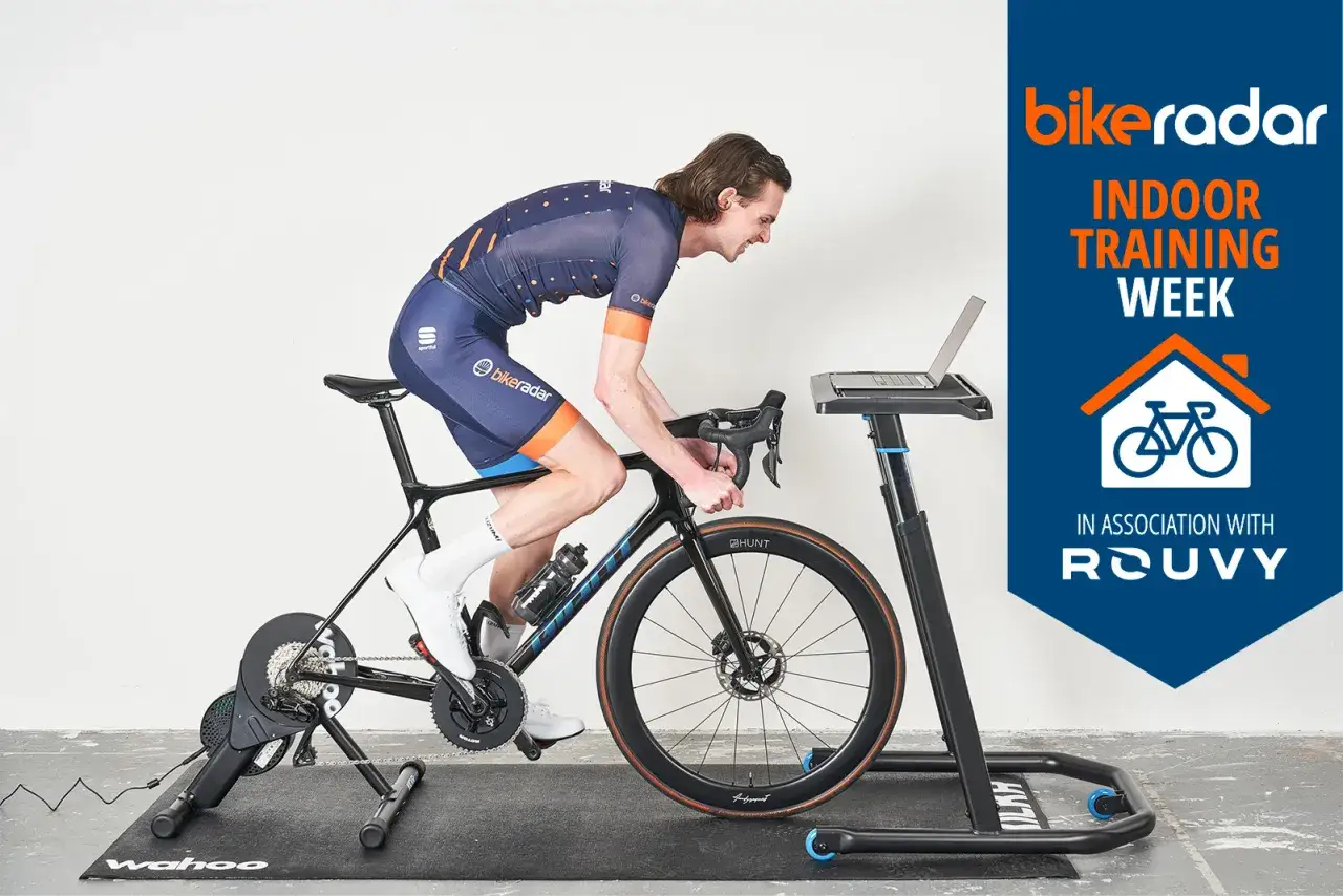 Kolarz w stroju sportowym odbywa intensywny trening na trenażerze rowerowym, z laptopem przed sobą.