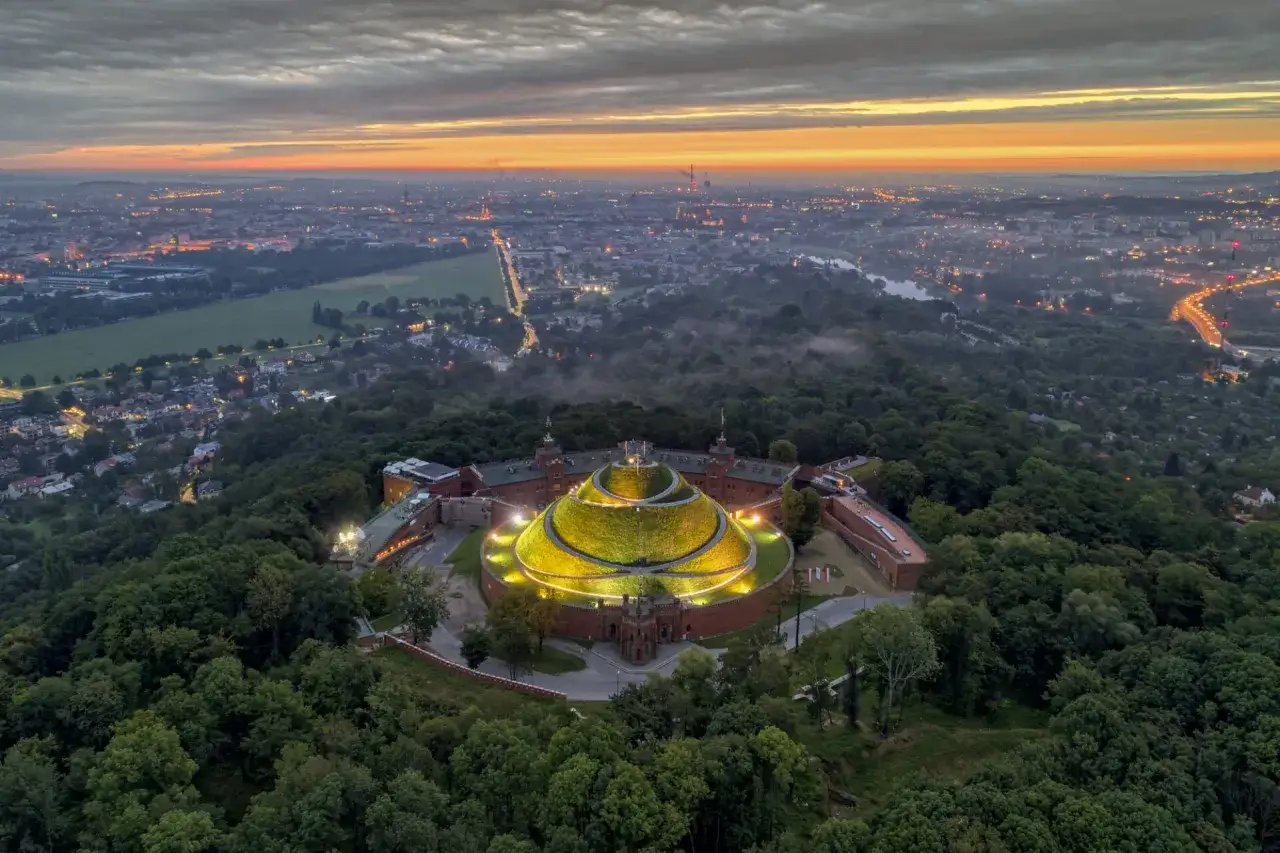 Krak&oacute;w o wschodzie słońca, z g&oacute;rującym nad lasem, oświetlonym Kopcem Kościuszki.