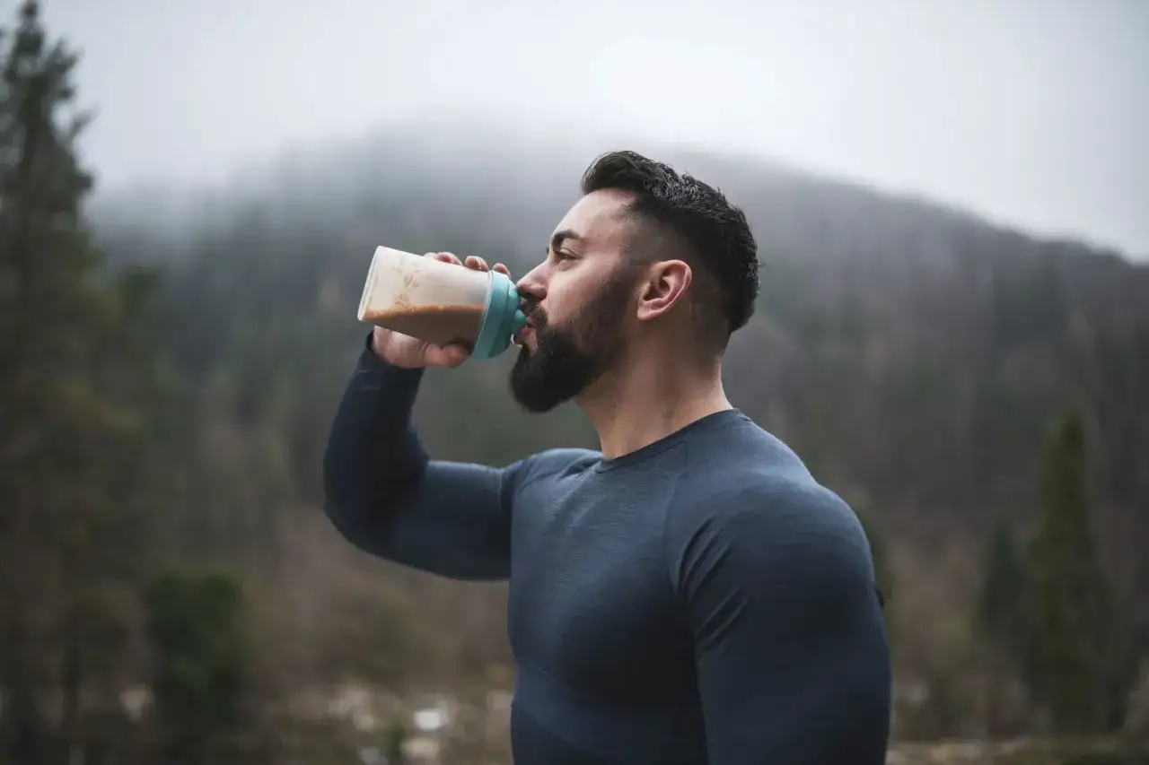Mann trinkt proteinshake vor oder nach dem Training in nebliger Berglandschaft.