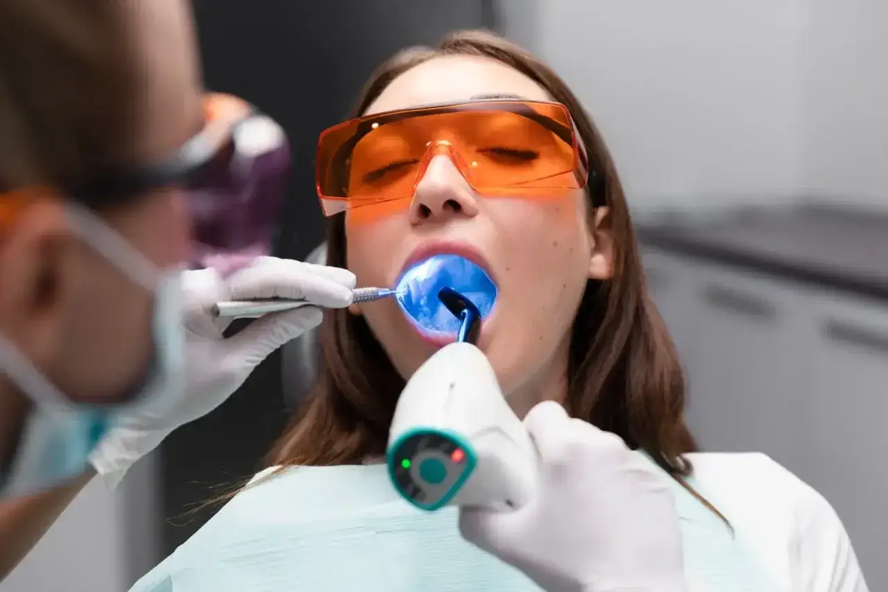 Pacjentka w okularach ochronnych poddaje się zabiegowi stomatologicznemu. Dentysta używa lampy polimeryzacyjnej, co jest jedną z metod fluoryzacji.