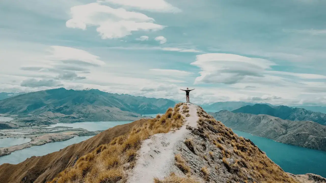 Ein Wanderer steht mit ausgebreiteten Armen auf einem Berggipfel, blickt auf eine weite Landschaft mit Seen und Bergen, ein Moment der Freiheit auf dem Te Araroa.