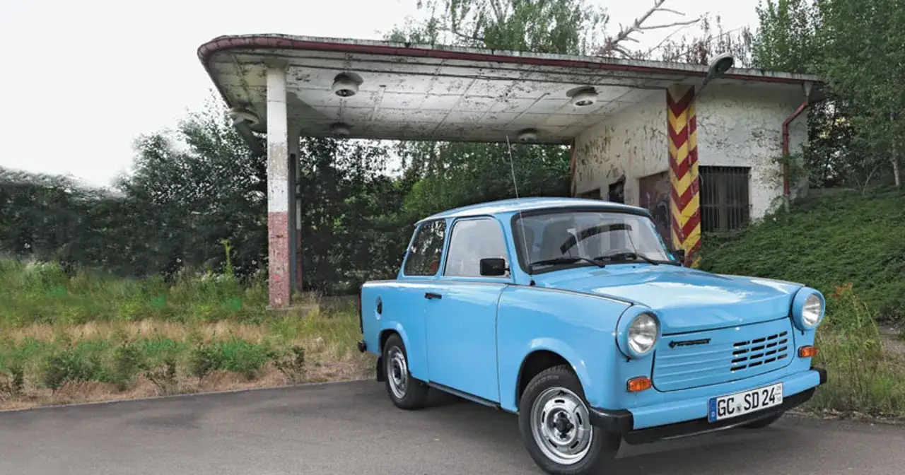 Niebieski trabant stoi przed opuszczoną stacją benzynową, otoczony bujną zielenią.