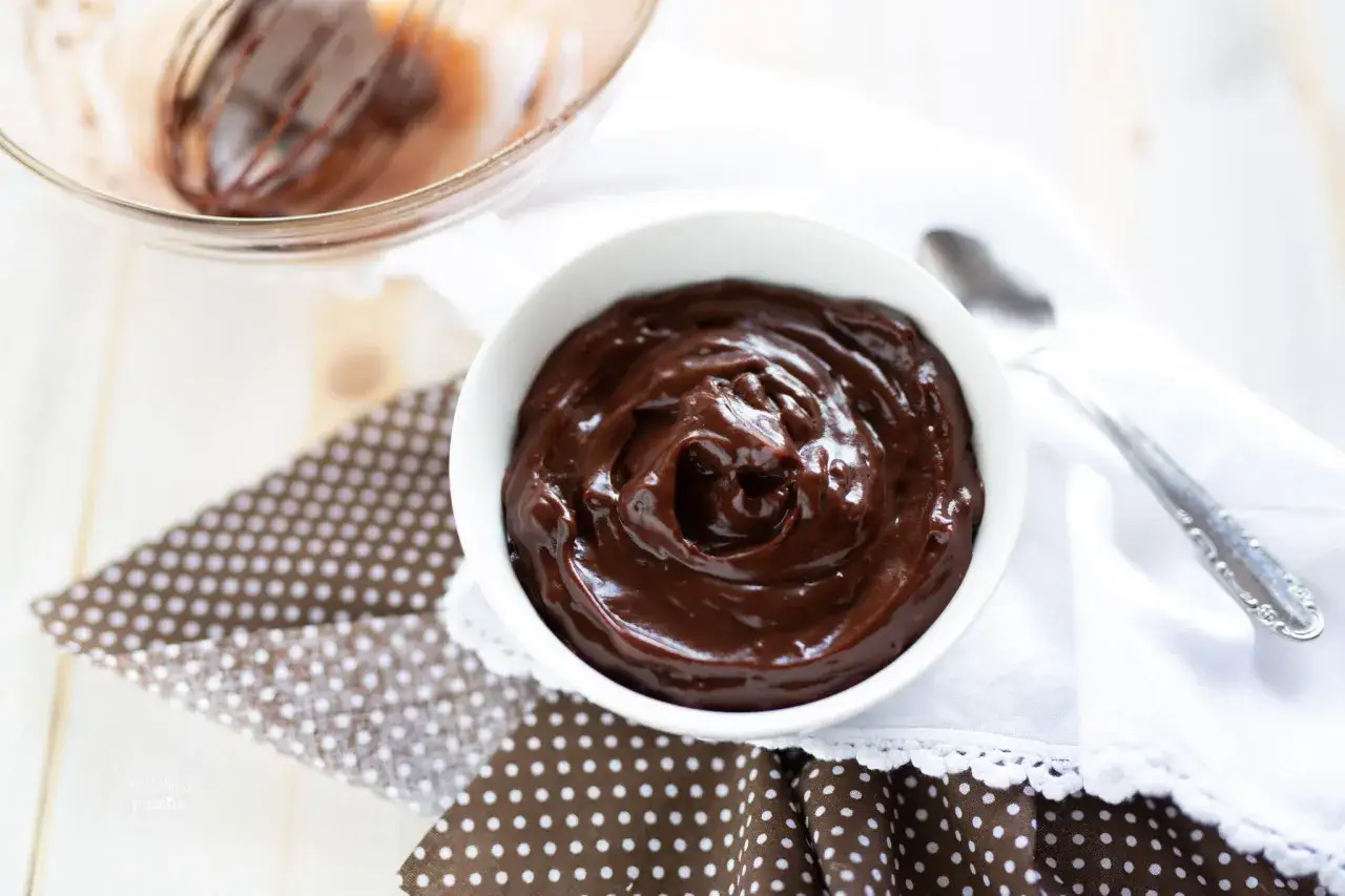 Una ciotola di lucida crema pasticcera al cioccolato, pronta per essere gustata. Vicino, una frusta in una ciotola di vetro.