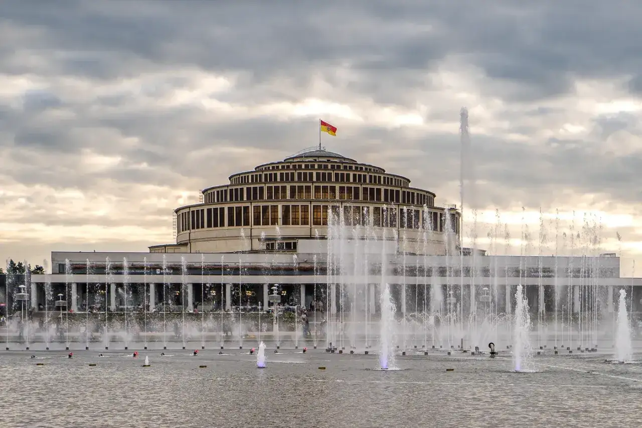Okrągły budynek Hali Stulecia we Wrocławiu z fontannami i flagą na szczycie. Iglica jest częścią tej monumentalnej budowli.