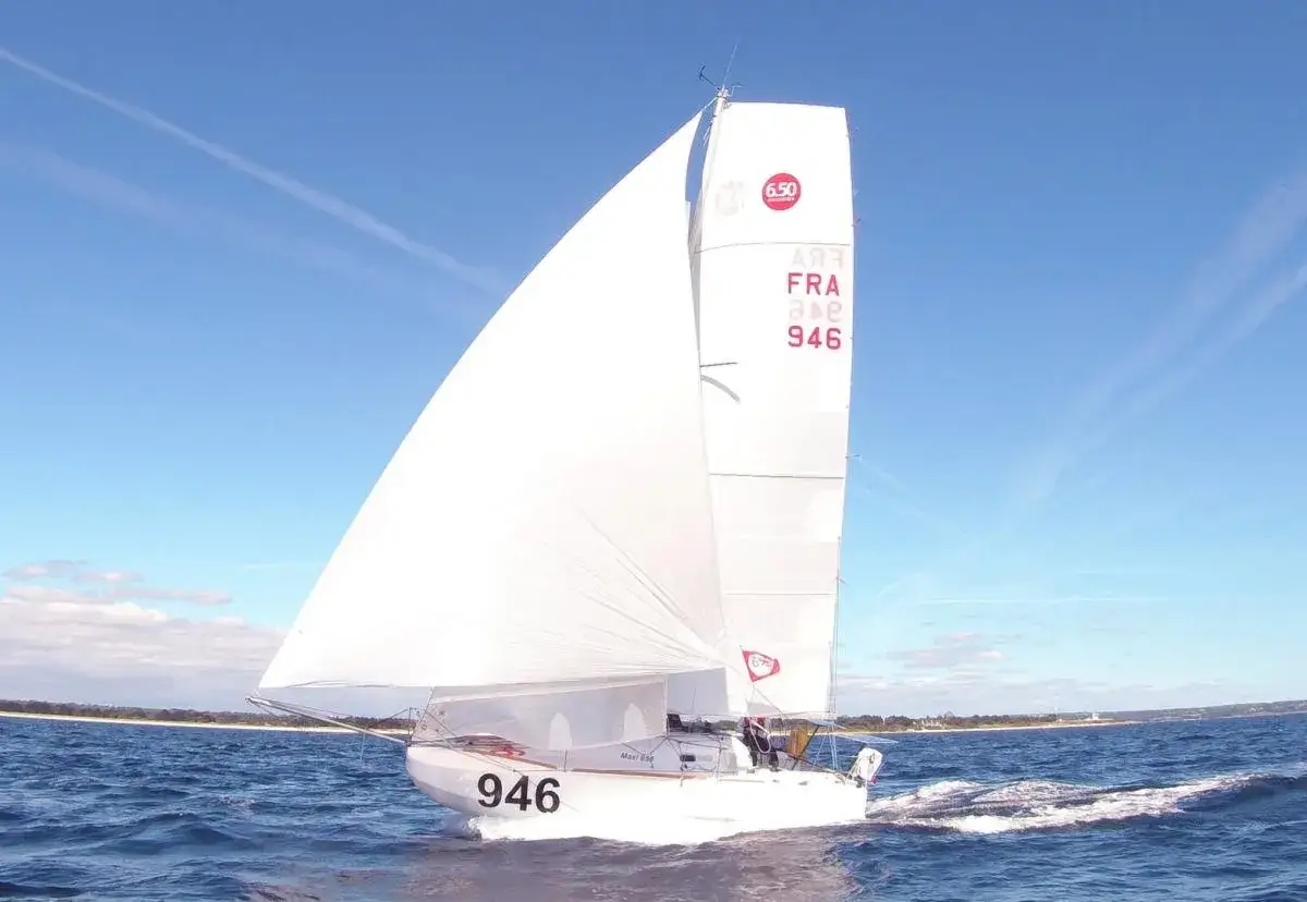 Un voilier de la classe mini, numéro 946, navigue sur une mer bleue sous un ciel clair.