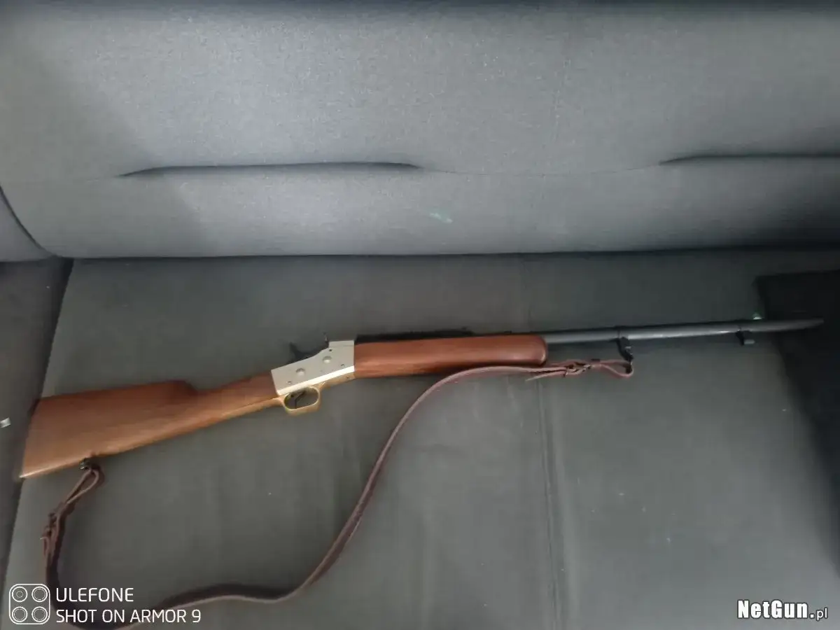 Karabin z drewnianą kolbą i metalowym zamkiem, model rolling block 50, leży na ciemnym tle.